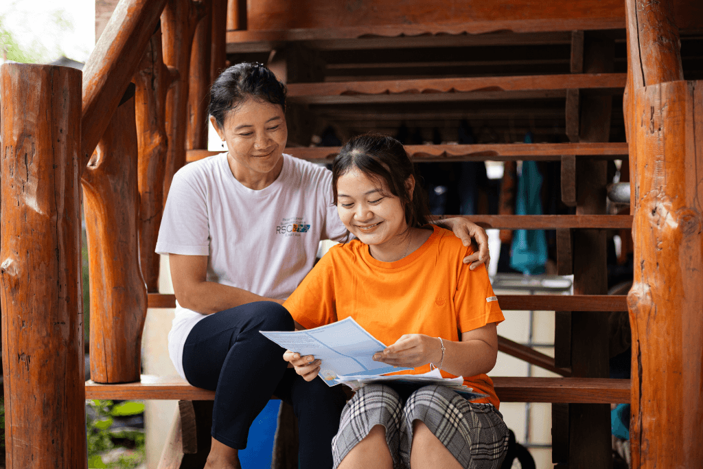 Young girl called Rosa sits with her mother on a set of wooden stairs as they smile reading a sponsors letter to them