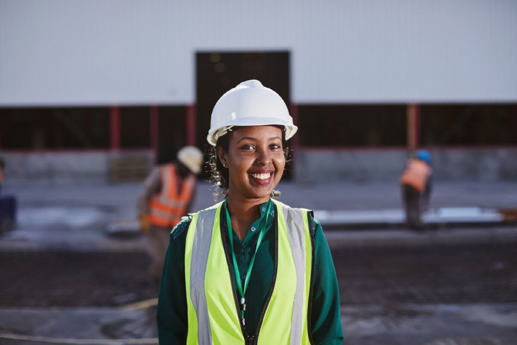 Bethelhem, in a yellow and grey safety vest, green shirt and white hard hat, is at the plant. She is standing in front of a large white building. There are workers in orange vests in the background.