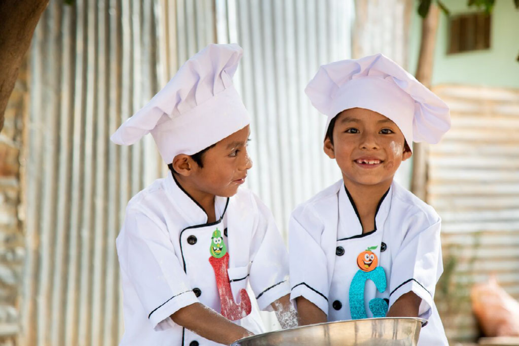 Gabriel (right) and Leonel, in white chef's hats and jackets, have their arms inside a silver bowl, playing with flour, which is also on their faces.
