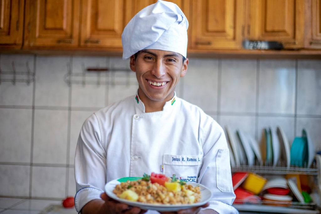Jesús is standing in the kitchen holding a plate of food. He is smiling and looking at the camera. Jesus is wearing a white chef's jacket and hat.