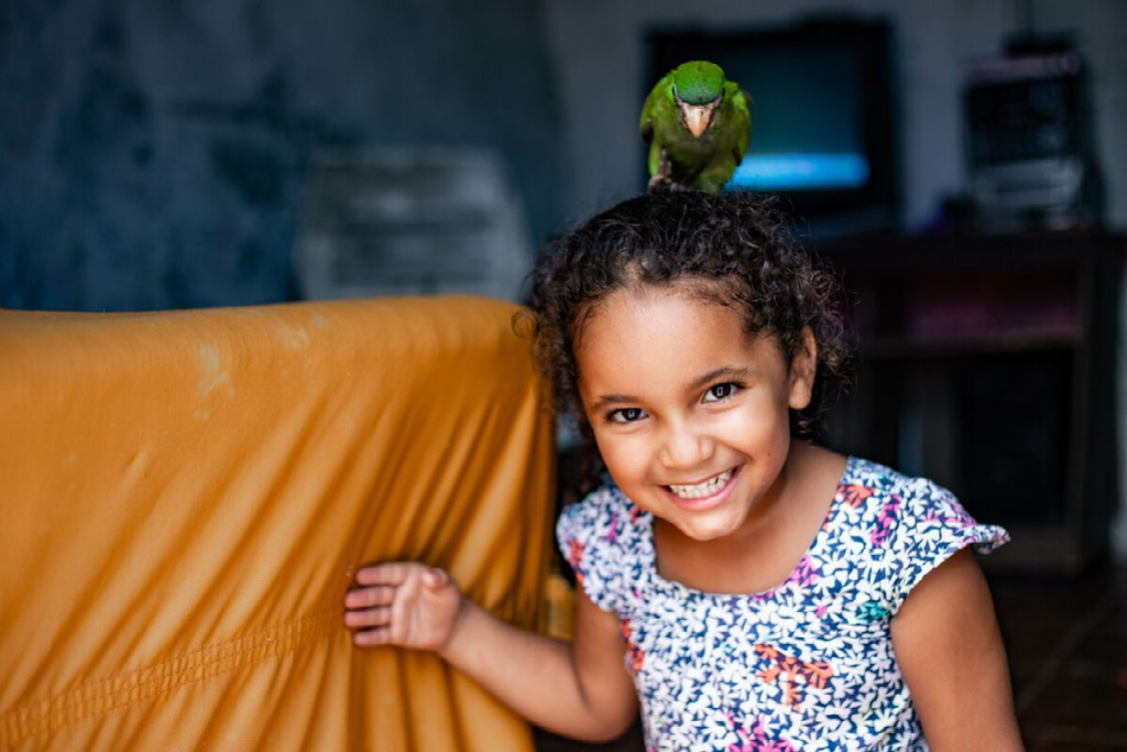 Tico, Maisa's aunt's green parrot, is on top of Maisa's head. They are inside Maisa's home next to a piece of furniture with an orange slipcover.