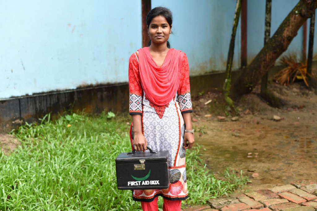 Moyna wears a red, white and black sari stands outside on a brick walkway holding a black first aid box. There are grass and a light blue building in the background.