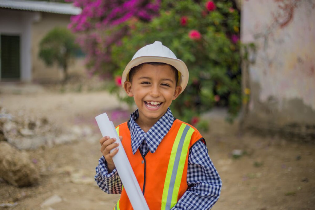 Santiago is wearing a civil engineer ‘s costume, an orange and yellow vest and a white hard hat. He is holding white rolls of paper, blueprints. There are building and flowering trees in the background.