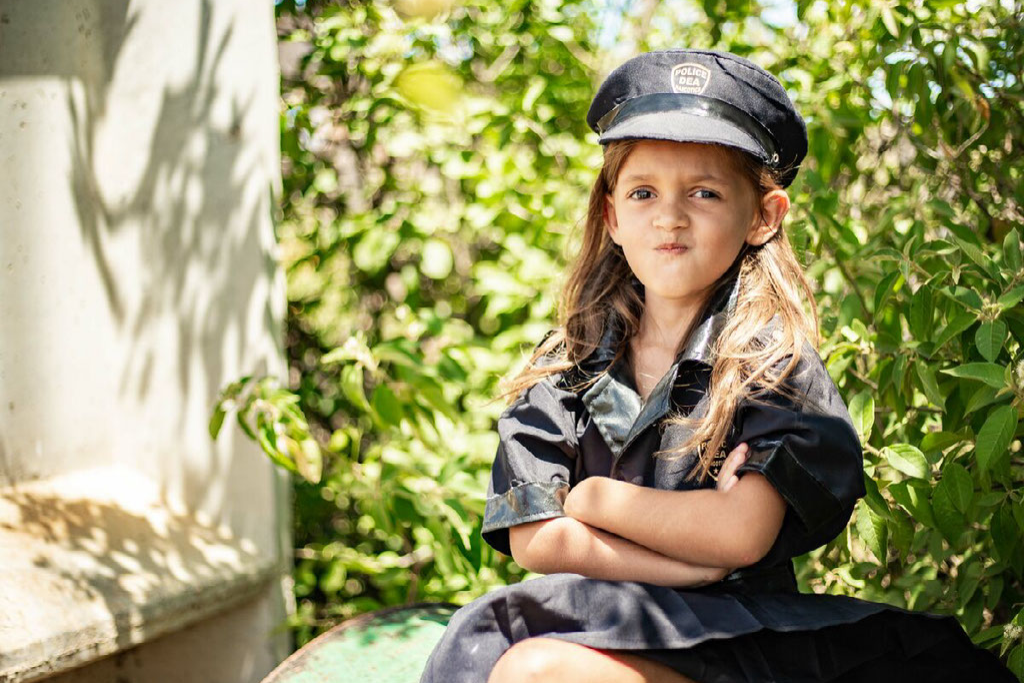 Tayla, in a black police officer uniform, is crossing her arms and faking a police officer face, a silly face. There are trees, leaves in the background.