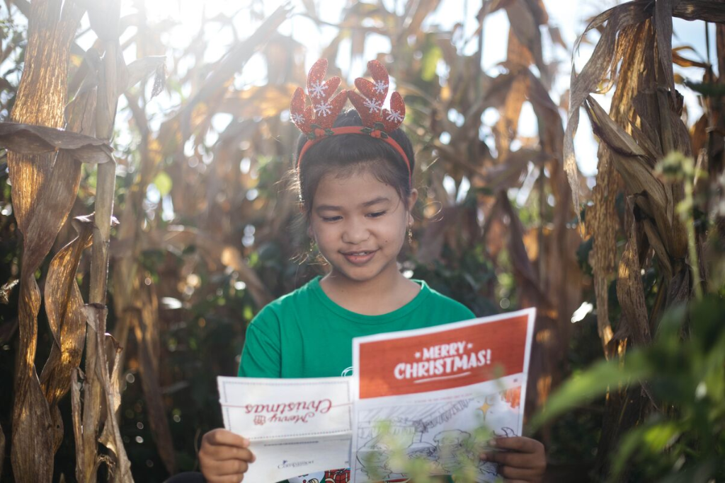 Ayu is wearing a green shirt and a headband with reindeer antlers. She is standing in a corn field and is reading a letter from her sponsor.