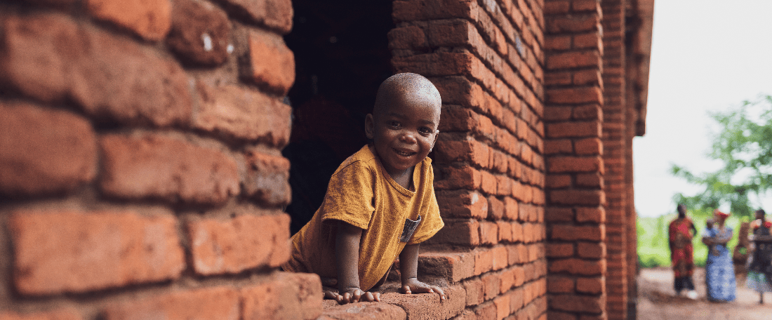A boy in a yellow shirt leans out of the window of a brick church building during a child registration event in Malawi.
