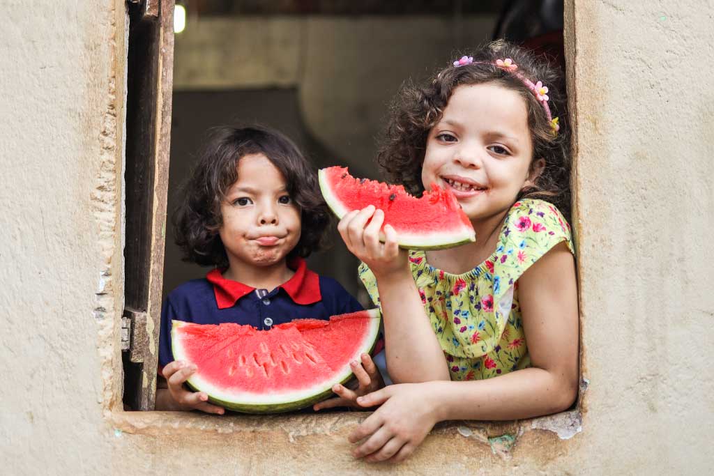 Siblings Tawany and Gustavo at a window eating watermelon