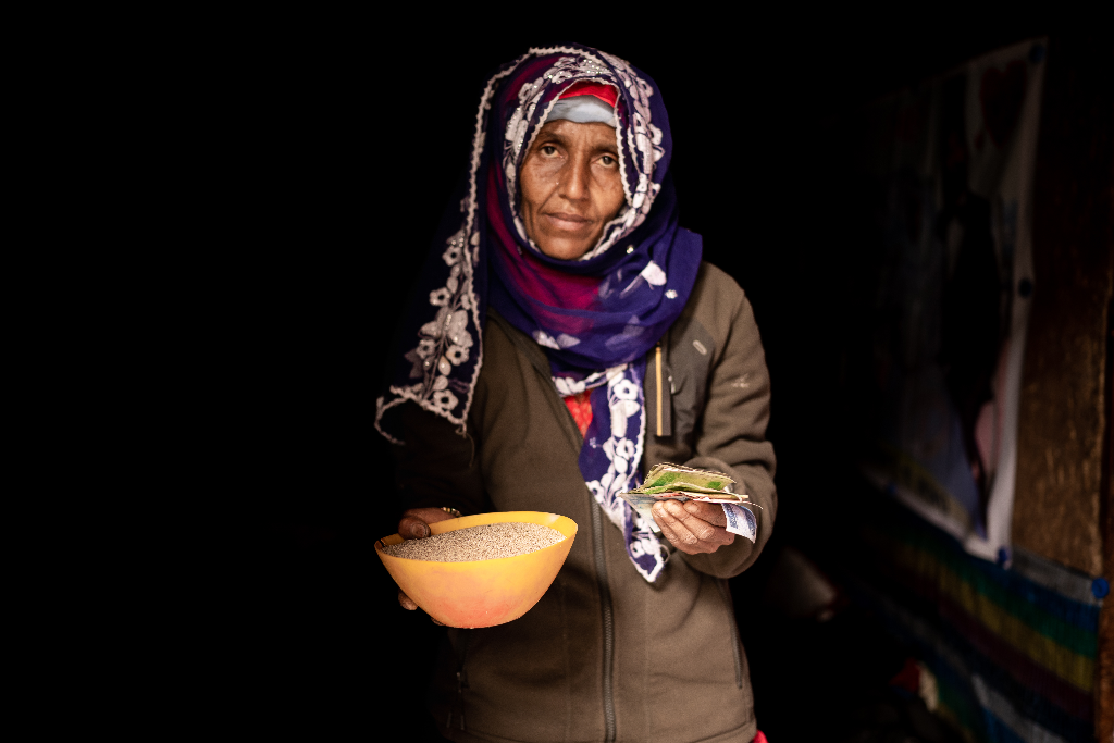 Fatuma, wearing a brown jacket and a colourful head covering, is holding a handful of money in one hand and a yellow bowl of grain in the other hand.